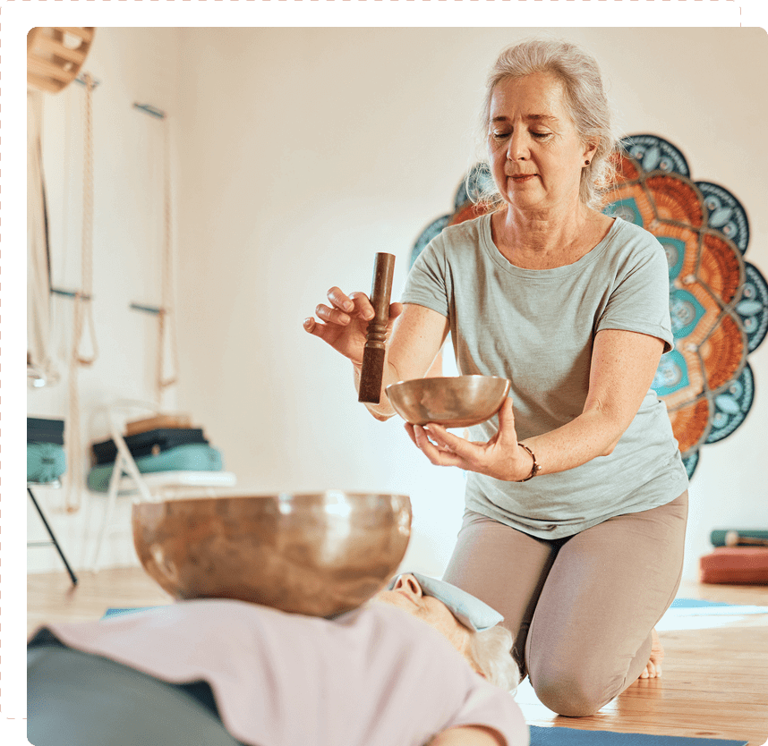 An older woman kneels on the floor holding a singing bowl and mallet near a person lying down in a yoga studio.