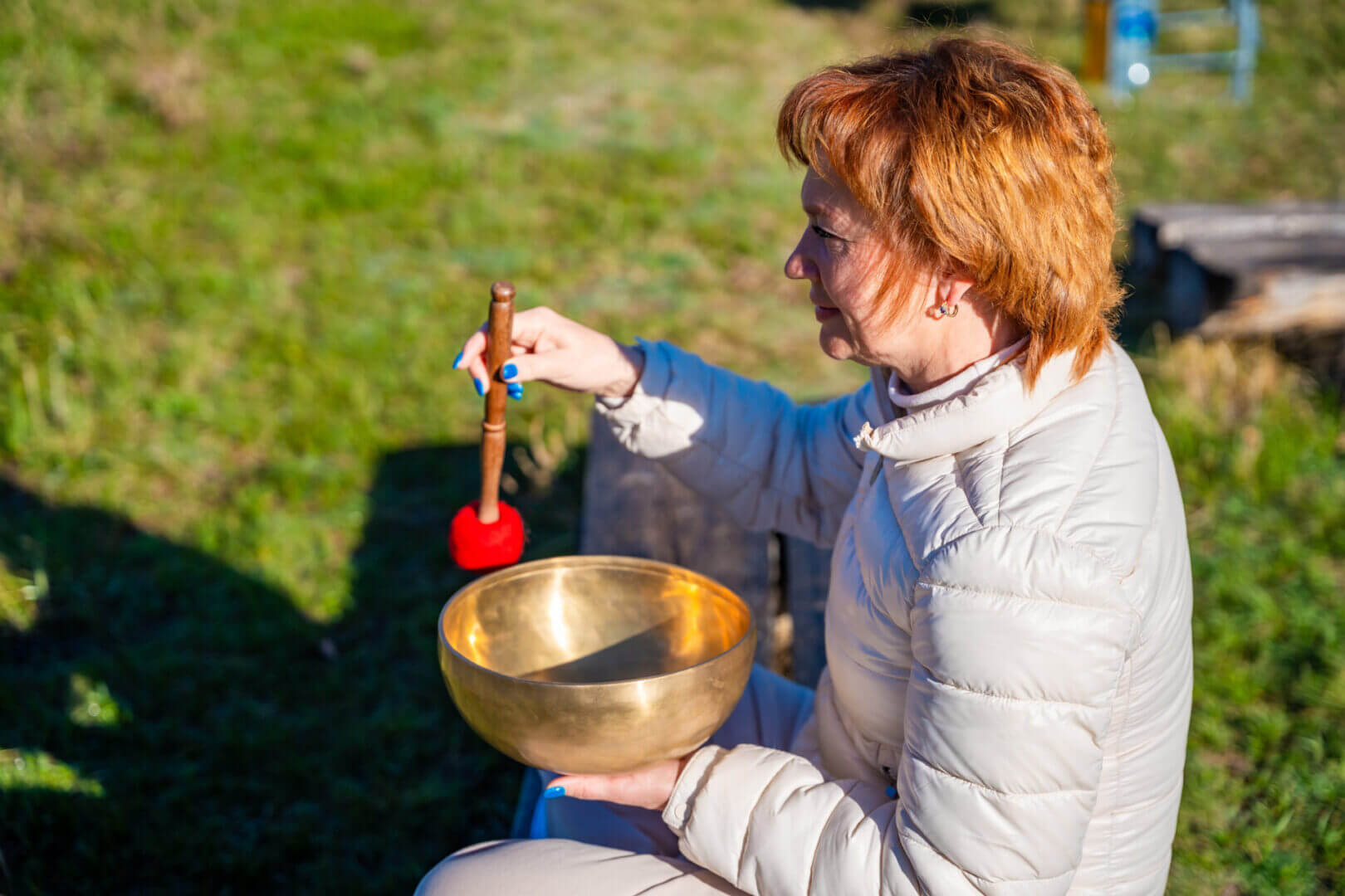 A person in a light jacket holds a brass singing bowl outdoors, using a mallet to play it, with green grass in the background.