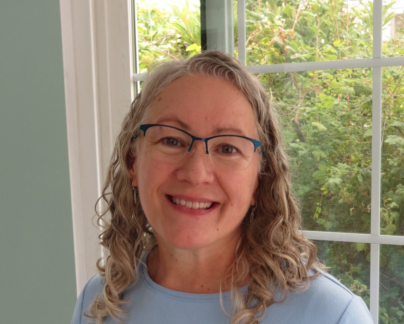 Smiling woman with glasses and curly hair standing by a window.