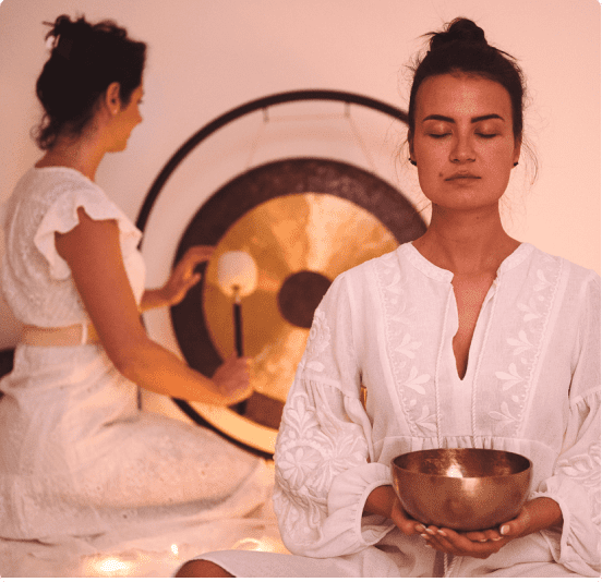 Two women meditating, one playing a gong and another holding a singing bowl.