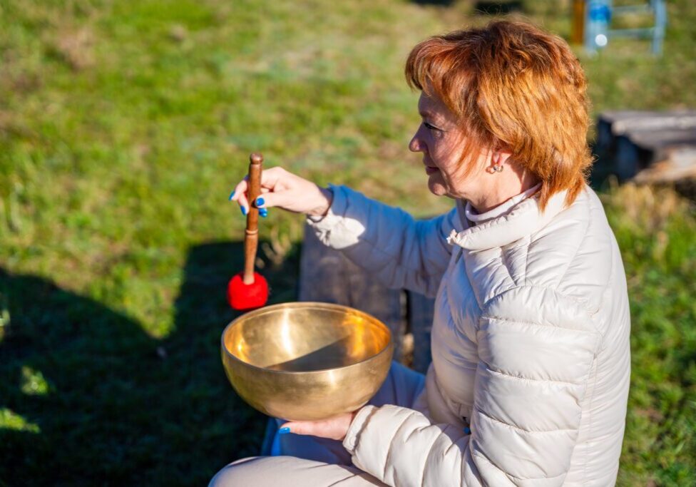 A person in a light jacket holds a brass singing bowl outdoors, using a mallet to play it, with green grass in the background.